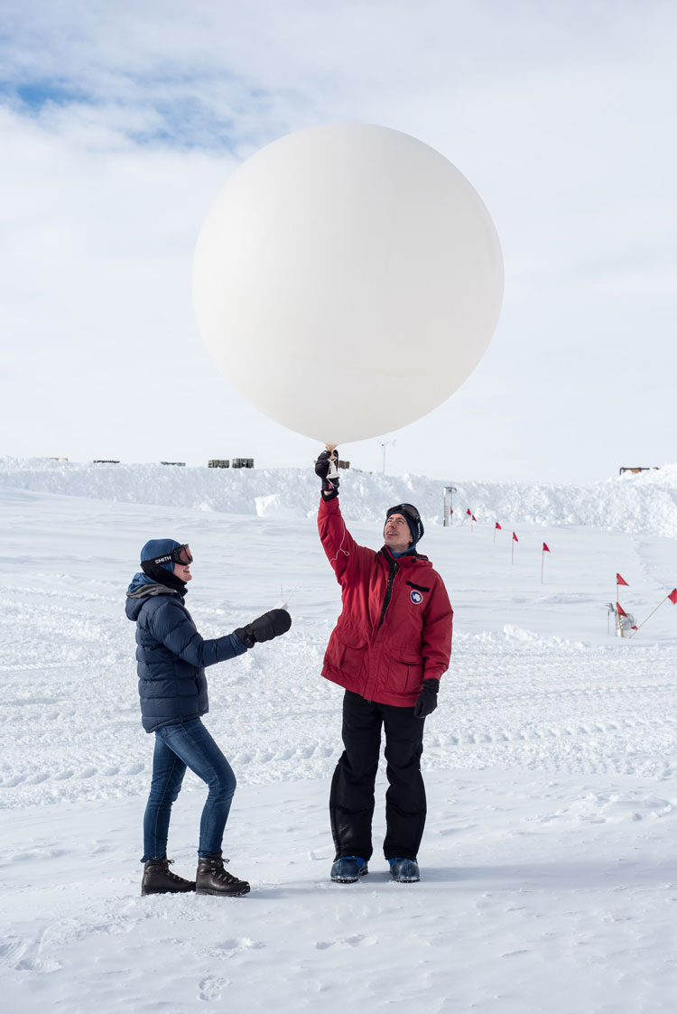 Two people standing on snow, holdingoverhead and about to release a large weather balloon.