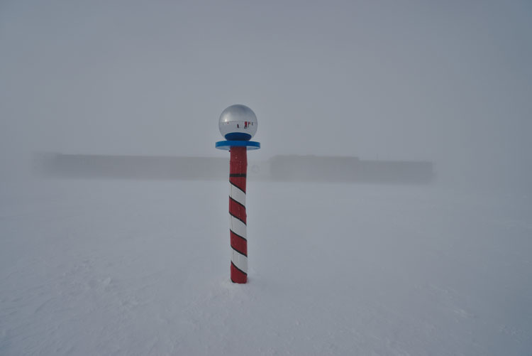 Ceremonial South Pole marker in foreground, station in background barely visible in whiteout