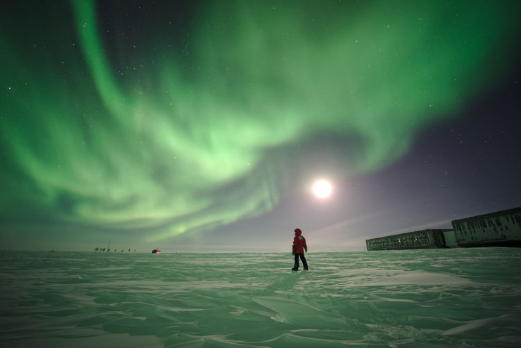 Lone person standing at a distance, swirling green auroras overhead, snow on surface reflecting green from above