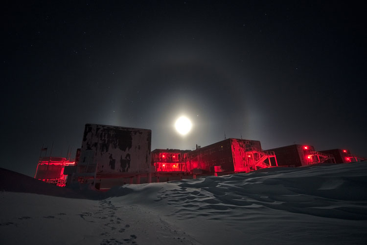 South Pole station bathed in red light with full moon and halo overhead.