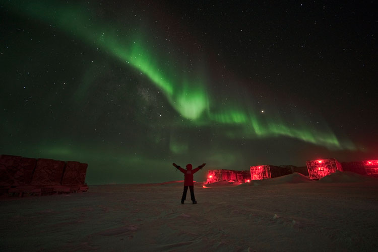 Person outside at the Pole, with arms raised up, and streak of bright auroras in sky above