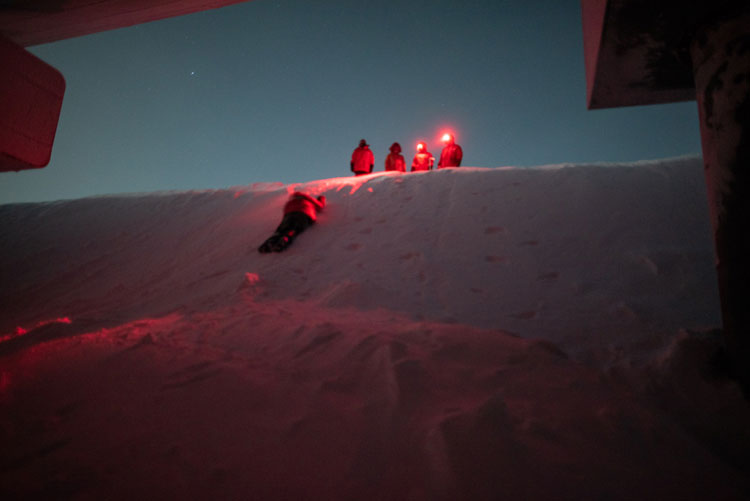 Person in red parka sliding down snow bank with others watching from above