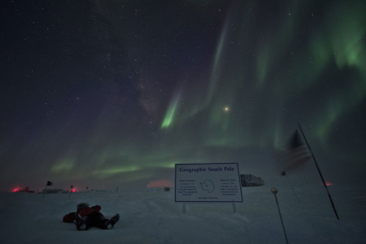 Person lying on snow at geographic South Pole sign, with auroras overhead