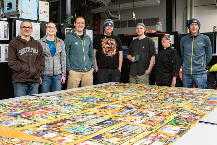 Seven people standing around table holding completed 18000 piece jigsaw puzzle