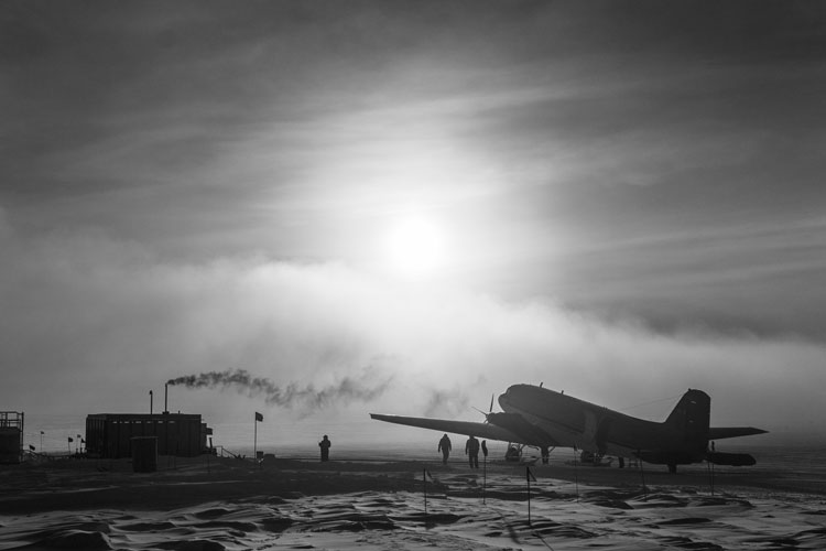 Black and white image of small jet parked at South Pole with a few people near it