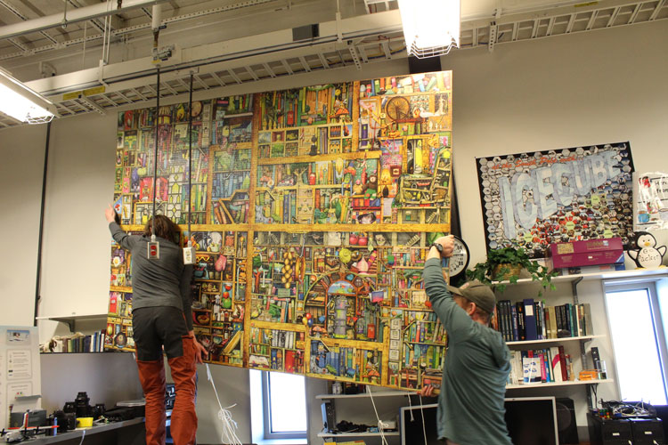 Two people hoisting up a very large jigsaw puzzle to be hung on wall.