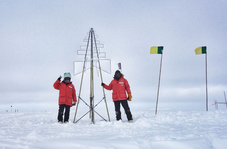 Two people in red parkas standing out on the snowon opposite sides of erected radio antenna