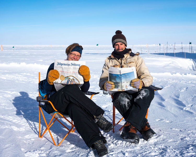 Two people sitting outside at the Pole in chairs, holding/reading newspapers
