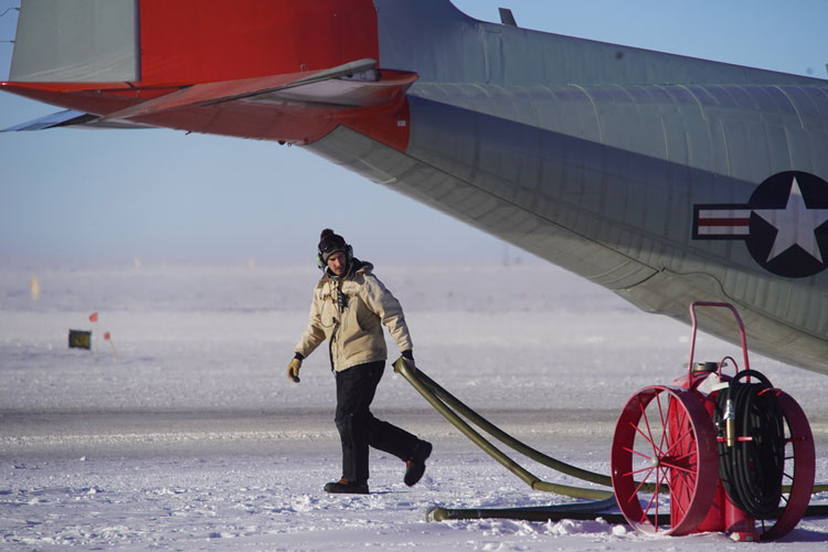 Person walking along, pulling long fuel hose underneath the tail of an aircraft