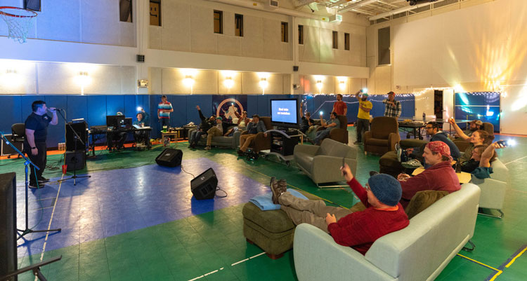 View of karaoke in gym, with person holding mic and group of spectators on surrounding sofas and chairs