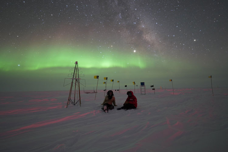 Two people seated outside on snow under auroras in sky