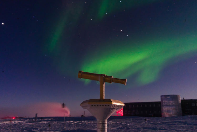 South Pole marker in foreground, auroras in sky above