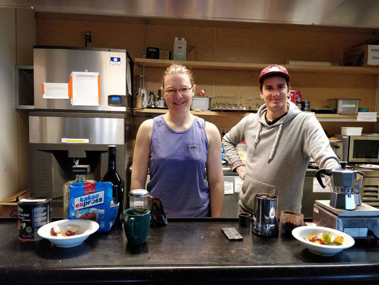 Two people smiling, behind counter laden with coffee items