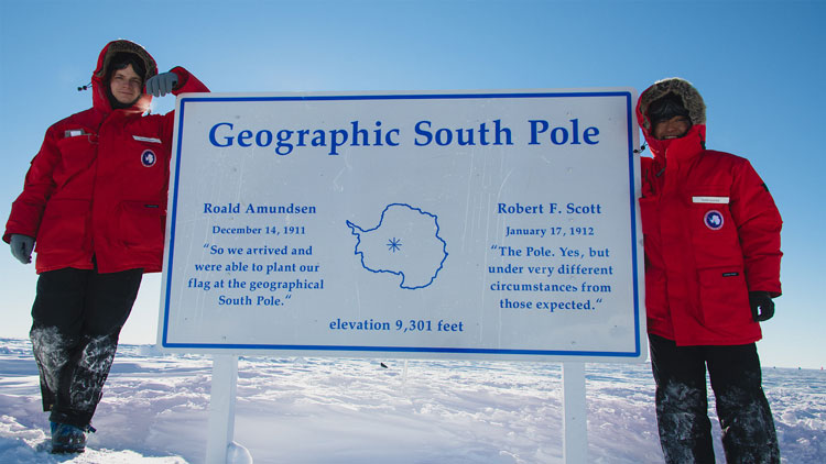 Two winterovers in red parkas flanking South Pole sign