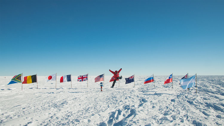 Person in red parka jumping into the air in distance at ceremonial South Pole, in front of line of flags