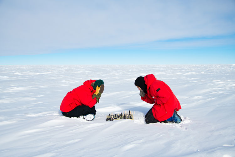 Side view, two people crouched outside at the south pole over a game of chess