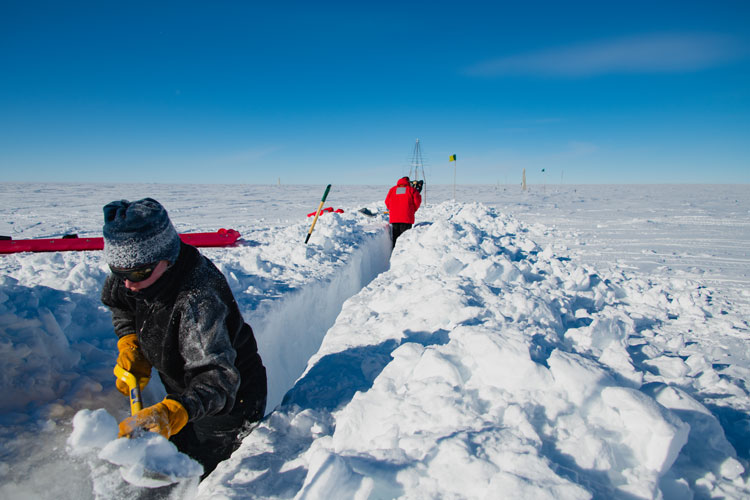 Two people, apart and facing away from each other, standing in and digging a deep trench in snow
