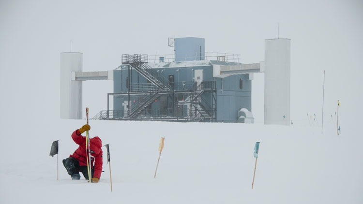 Person in red parka crouching outside taking snow measurements at the South Pole