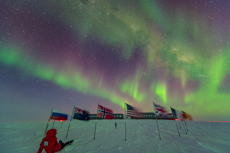 Person in red parka seated at ceremonial South Pole, facing away, with auroras in sky
