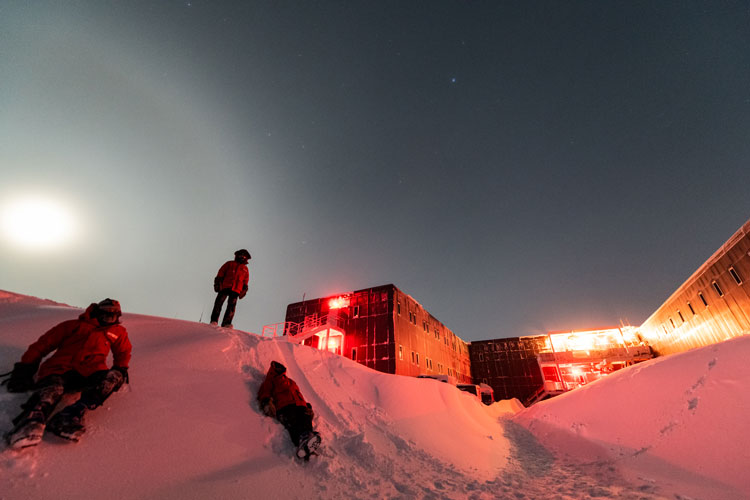 Three people in red parkas on snow bank at South Pole unde bright moon