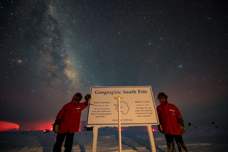 Two people posing under starry sky at geographic South Pole marker