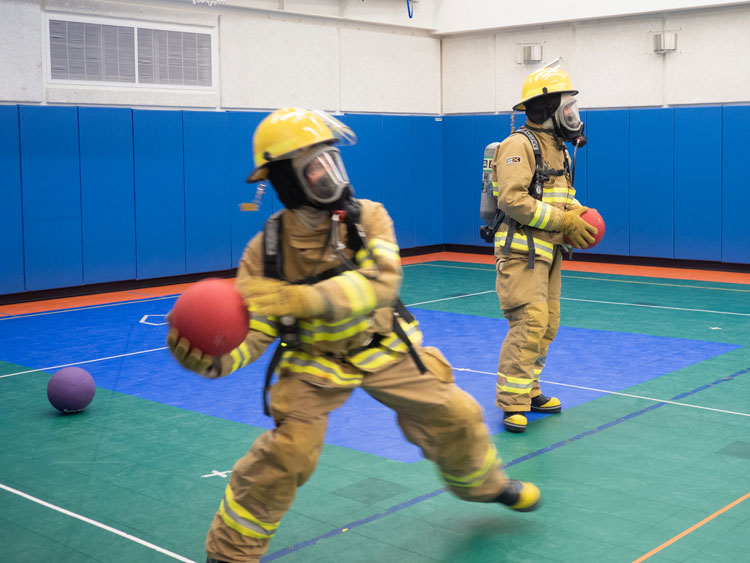 Two people in fire gear playing dodgeball in gym.