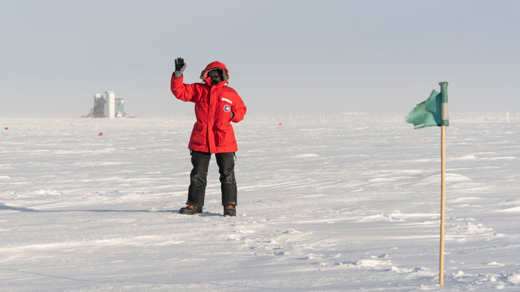 Person in red parka waving, with the IceCube Lab in the distance.