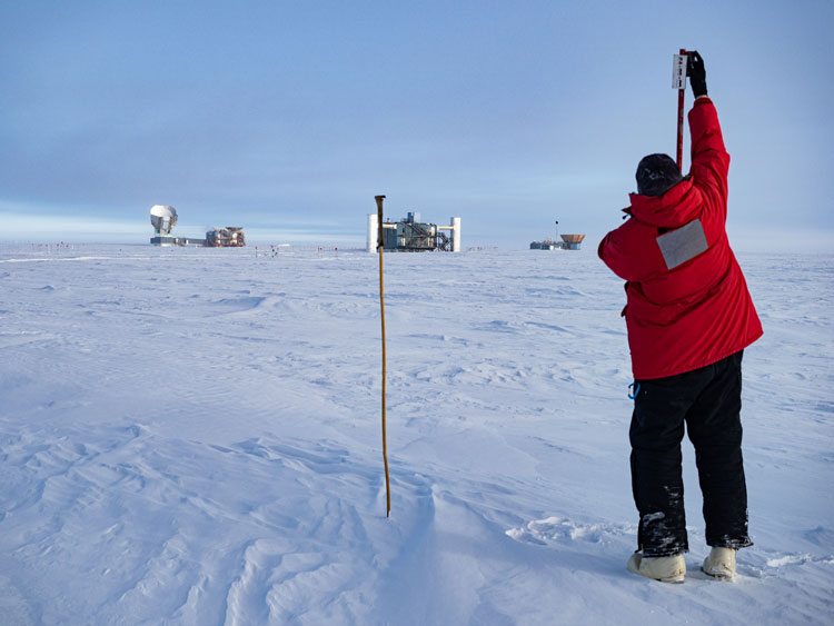 Winterover holding up measuring device, with IceCube Lab in background.