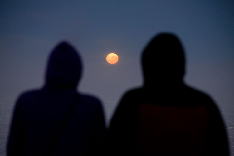 Blurry images of shadowed behinds of two hooded individuals facing a full moon, shown between them.