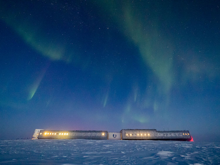 South Pole station on horizon, with light showing in some of the windows, and light auroras overhead.