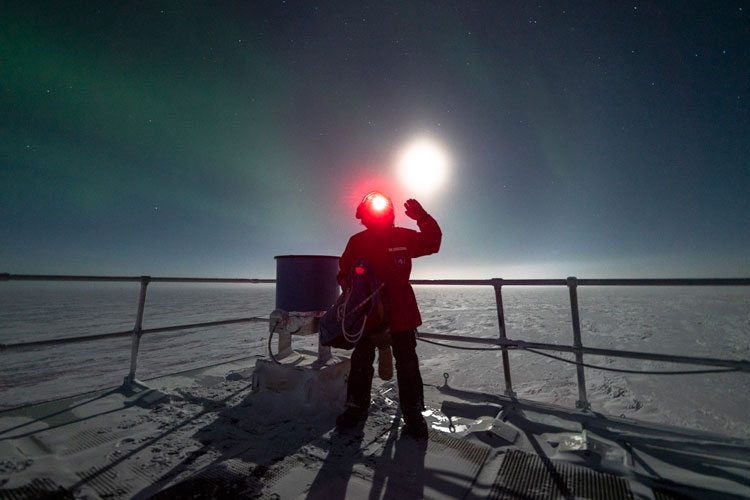 Person wearing red headlamp facing camera and waving, standing next to IceAct telescope, with bright moon just above their head.