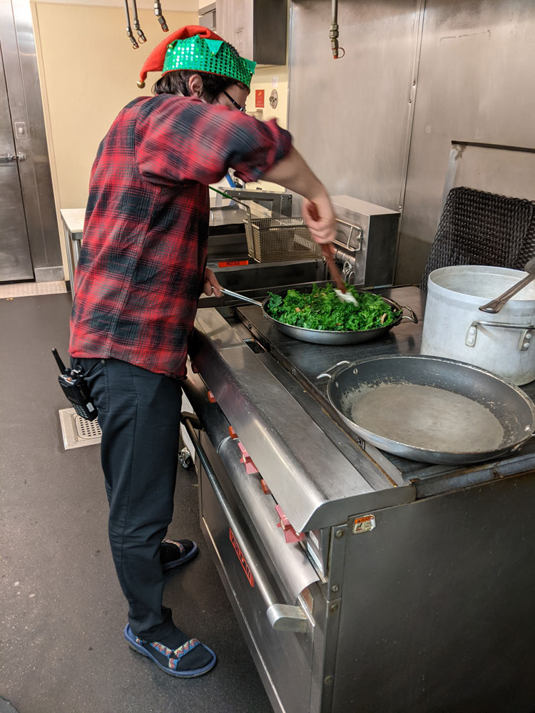 Person seen from side, standing at industrial stove and stirring a pan of greens.