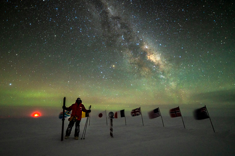 Winterover standing with ski poles at the ceremonial Pole with Milky Way overhead.