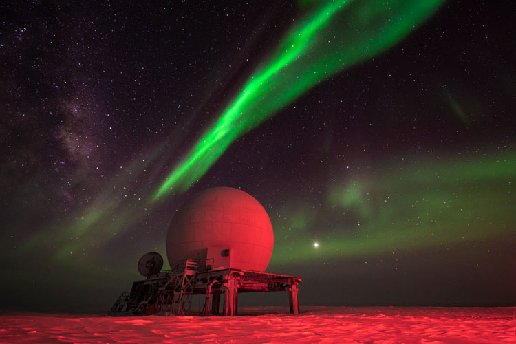 Satellite dome lit in red light, with streak of bright green aurora overhead.