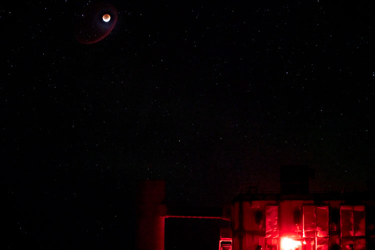 Super blood moon high in dark sky over IceCube Lab, lit in red.