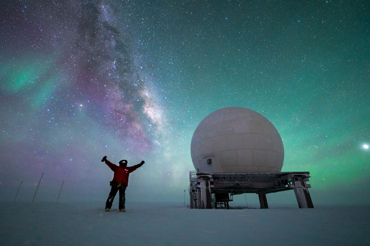 Sky full of auroras and Milky Way, person standing with arms up near a satellite dome.