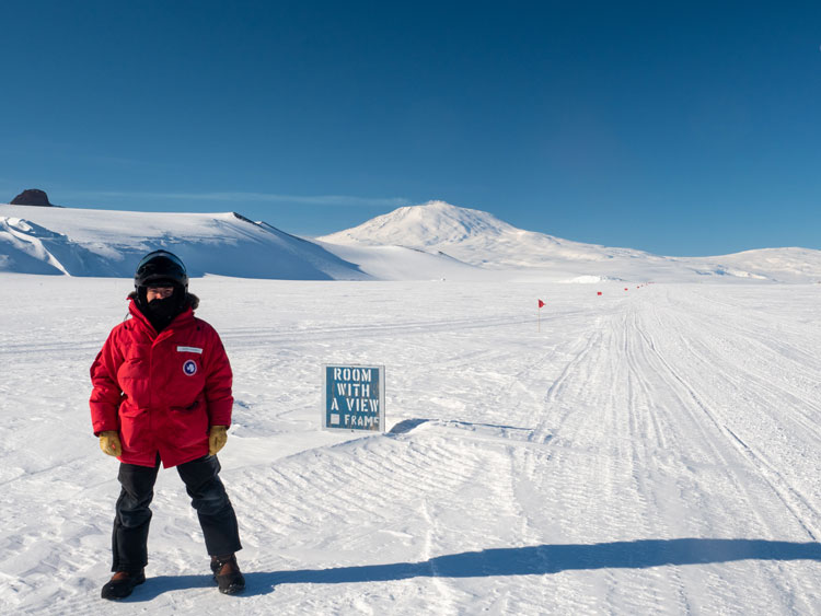 Person standing next to sign "Room With A View" out on snowy plateau, Mount Erebus in background.