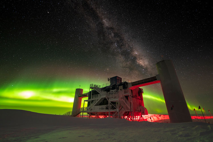 IceCube Lab backlit by low green auroras, with Milky Way overhead.