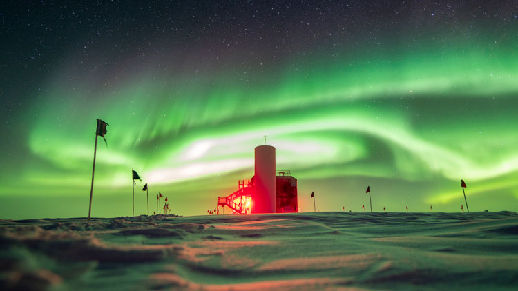 Side view of IceCube Lab with sky full of swirling green auroras.