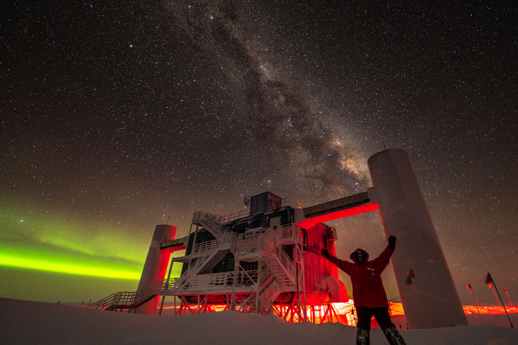 Person with arms up in front of IceCube Lab backlit by low green auroras, with Milky Way overhead.
