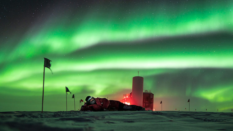 Side view of IceCube Lab with person lying on side on the ice and sky full of swirling green auroras.