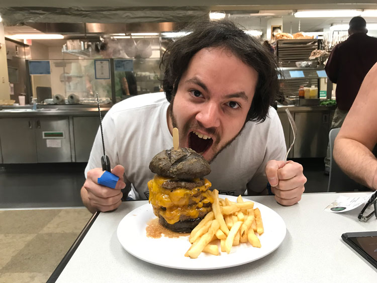 Josh with huge, stacked cheeseburger in front of him, and with mouth wide open as if about to devour it. 