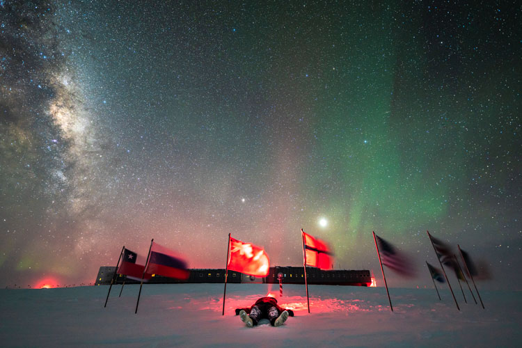 Winterover Martin lying on ground among flags at ceremonial Pole under starry skies, with view of station behind.