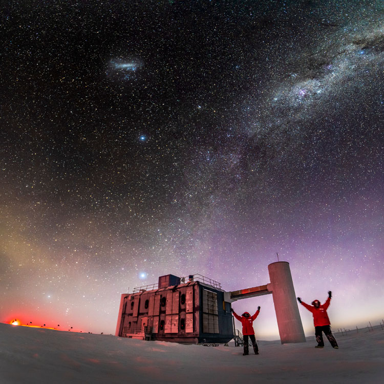 Winterovers Martin and Josh taking selfi outside the IceCube Lab, with arms up, stars in sky and sunlight along horizon.