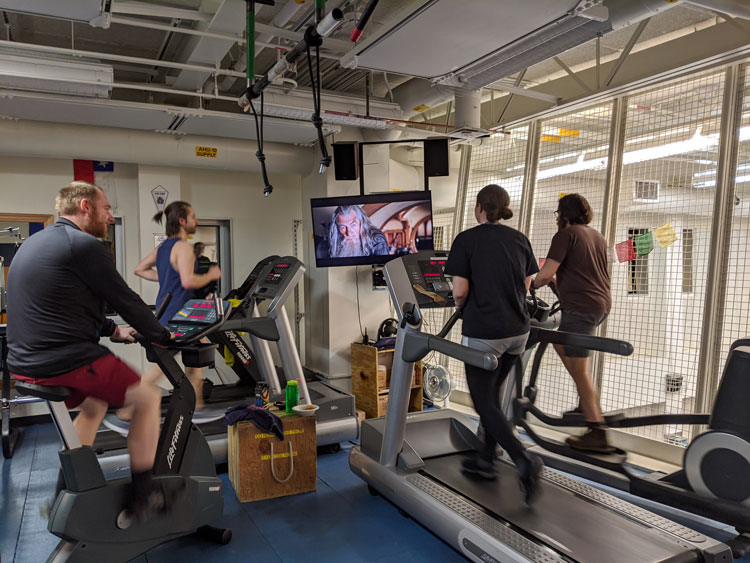 People in gym watching a monitor while exercising on treadmills and exercise bikes.