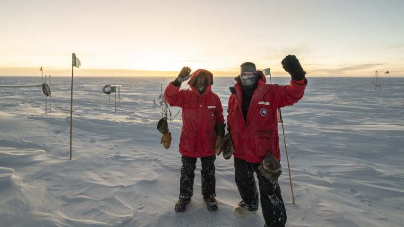 Two winterovers outside in red parkas waving at camera.