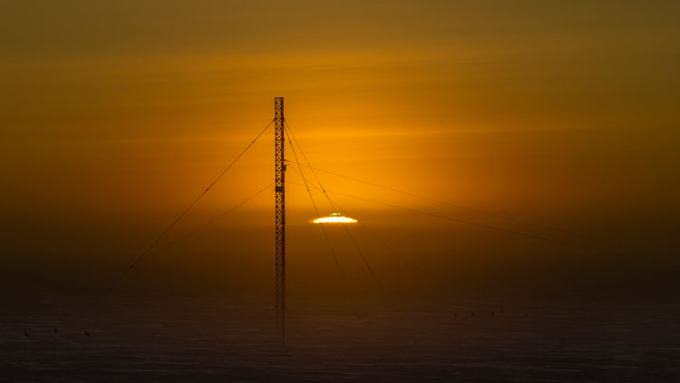 Sun rising at the South Pole, just at horizon with green flash visible, communications tower in foreground.