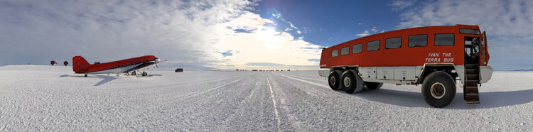 Wide-angle view of skiway at the South Pole, with a small red Basler plane parked on the left and a red shuttle bus parked off to the right.