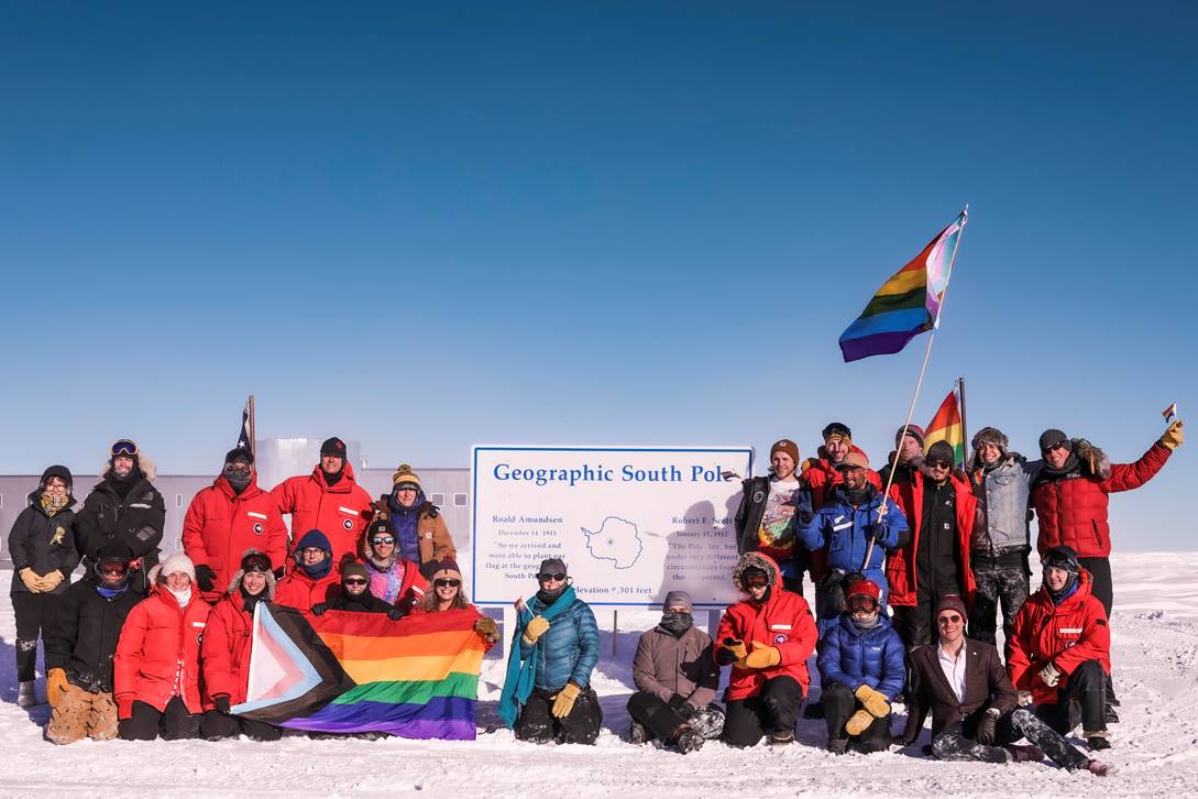 Group photo gathered around the geographic South Pole sign with pride flags.