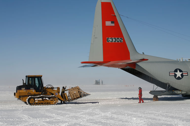 Side view of snow tractor with cargo load behind the tail of an LC-130, with a glimpse of the IceCube Lab in the distance between the tractor and the plane.